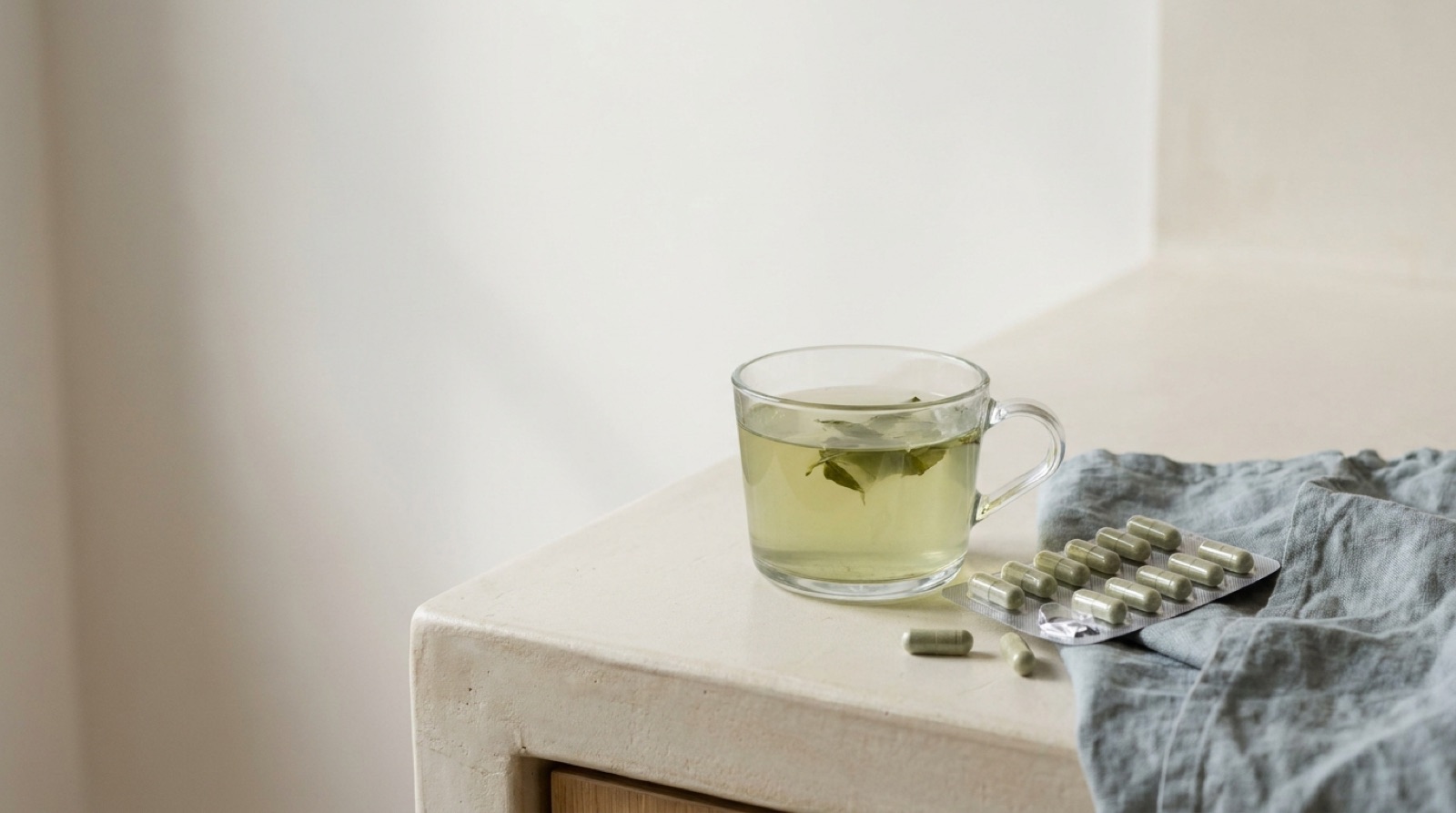 Ein Glas grüner Tee neben einer geöffneten Blisterpackung mit Kapseln auf einer hellen Ablage.